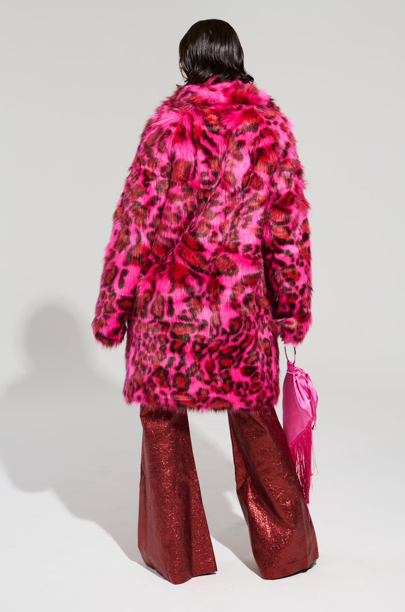 Dressed in the CHEETAH DYNASTY PINK MULTI FAUX FUR JACKET, a person stands with their back turned, pairing it with sparkling red flared pants. They hold a pink fringed handbag, all set against a plain gray backdrop that accentuates this bold winter fashion statement.