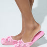 A close-up of a person's lower legs shows them wearing AZALEA WANG CHERINA pink flats with embellishments. These stylish slip-ons feature a mesh design decorated with floral appliques against a light, neutral background.