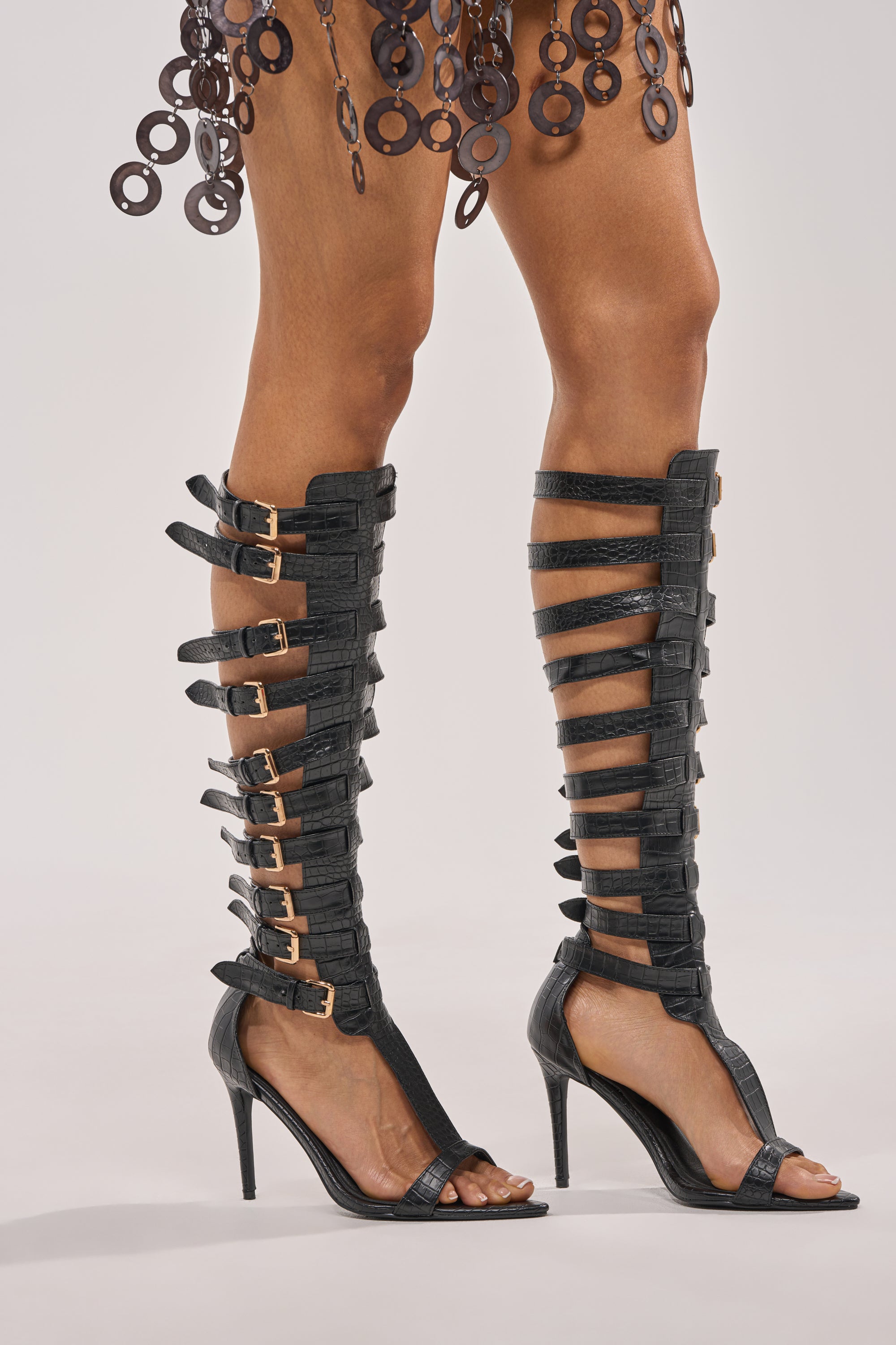 A person wears AZALEA WANG CHERLLI BLACK CROC OVER THE KNEE SANDALS with multiple gold-buckled straps, faux leather stiletto heels, and a skirt with large circular decorations, all set against a light background. 1