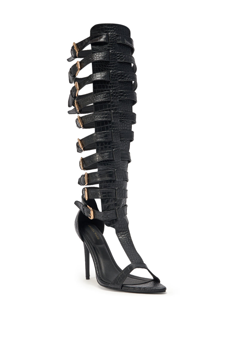 The AZALEA WANG CHERLLI BLACK CROC OVER THE KNEE SANDAL features a knee-high, black croc-embossed design with multiple horizontal straps, gold buckles, a stiletto heel, and open toe faux leather against a plain white background. 4
