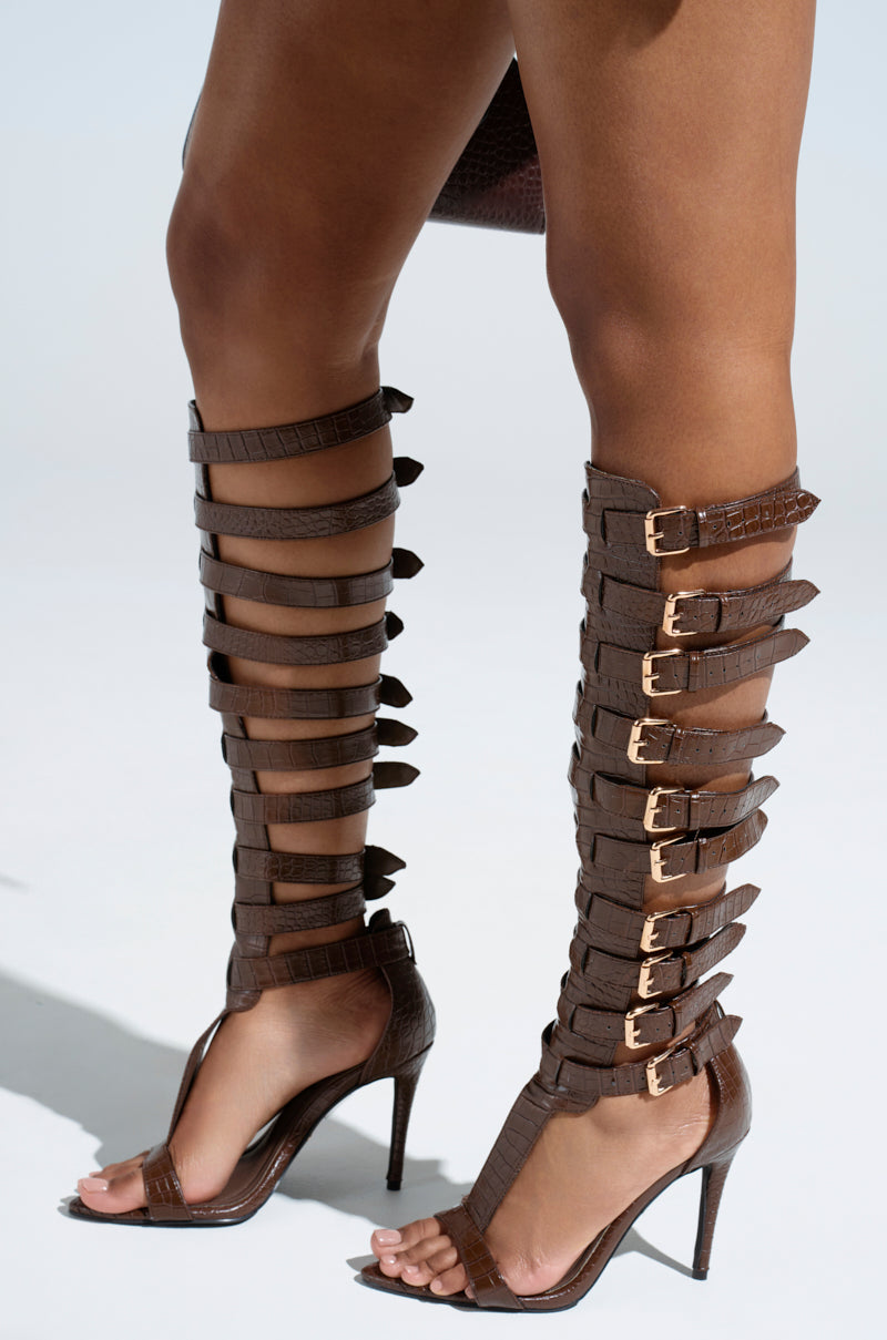 Close-up of AZALEA WANG CHERELLI BROWN CROC OVER THE KNEE SANDAL on a person. The knee-high gladiator features multiple straps and buckles crafted from croc-embossed leather, with high stiletto heels against a plain, light background.