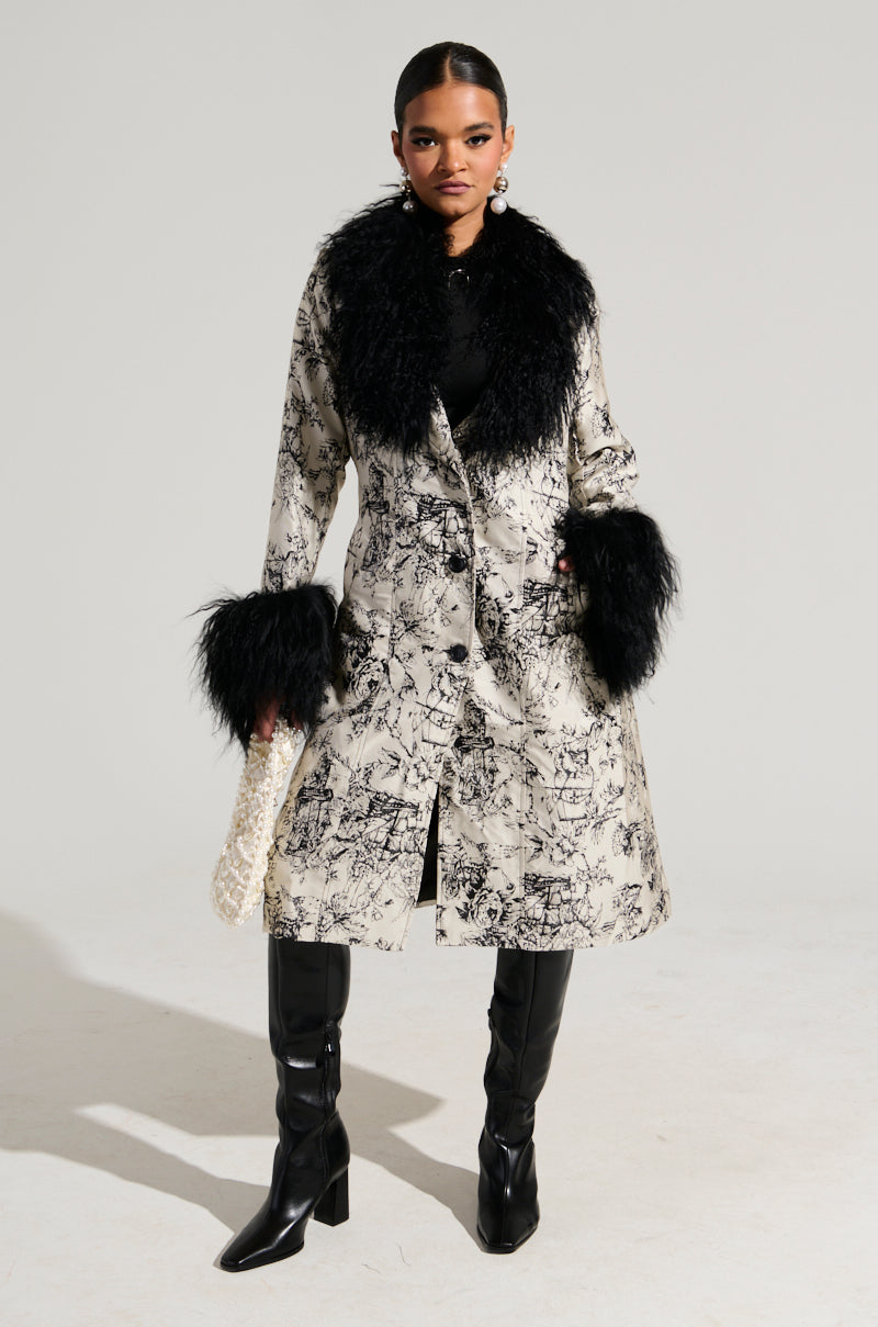 A person stands confidently in a chic TALULAH REMOVABLE FUR TRIM TRENCH IN IVORY MULTI, a 70's-inspired coat with striking black and white patterns. The stylish design, featuring detachable faux mohair fur trim and perfectly paired with black boots and dangling earrings, radiates fashionable elegance against a plain backdrop.