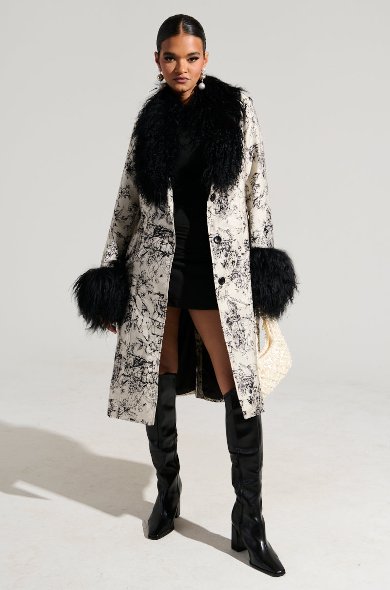 A person wearing a stylish black dress paired with the TALULAH REMOVABLE FUR TRIM TRENCH IN IVORY MULTI, which showcases 70's-inspired faux mohair fur trim on the collar and cuffs, stands confidently in knee-high black boots and pearl earrings against a plain background.
