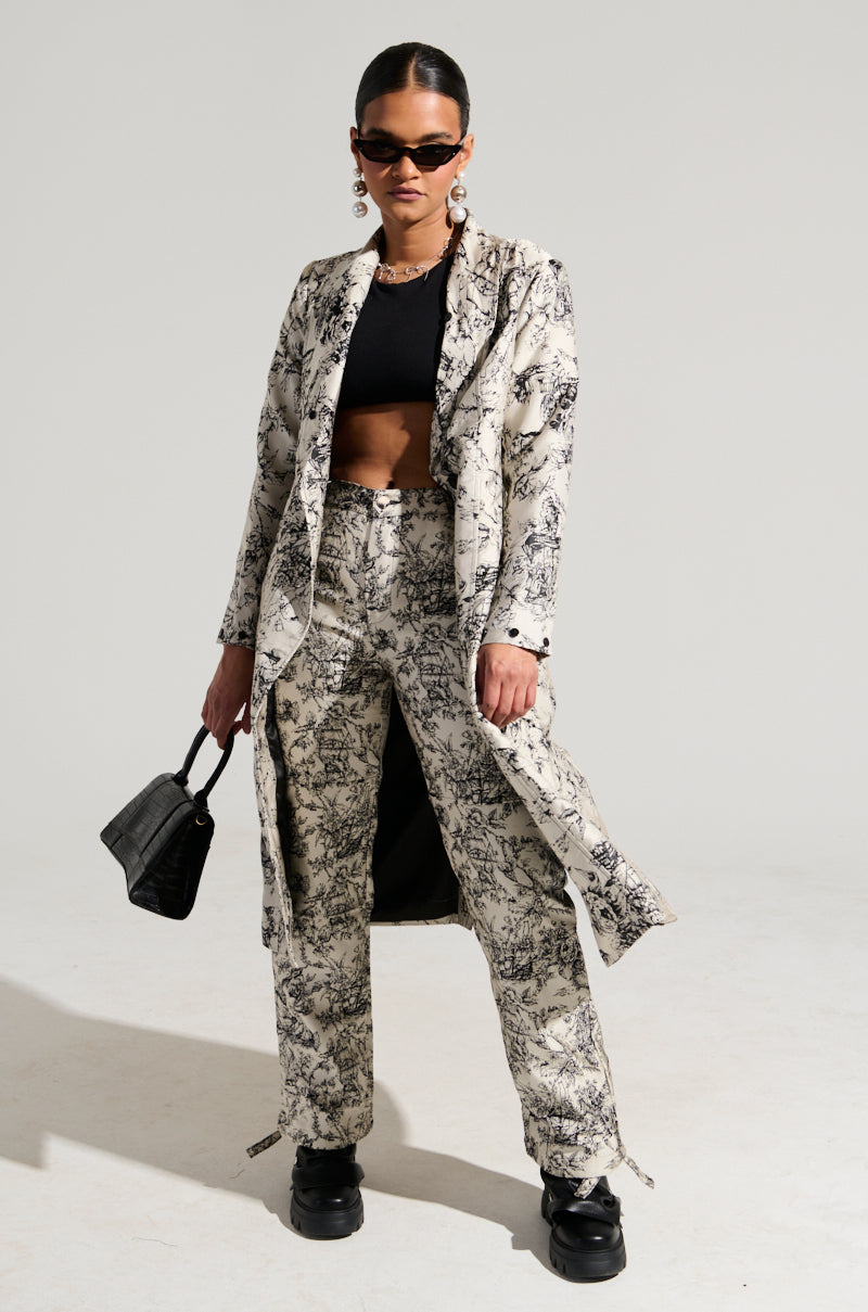 Wearing the TALULAH Removable Fur Trim Trench in Ivory Multi along with matching pants, a person pairs their 70’s-inspired look with a black crop top and chunky shoes. Posing confidently, they hold a chic black handbag and sport dark sunglasses, their hair pulled back neatly.