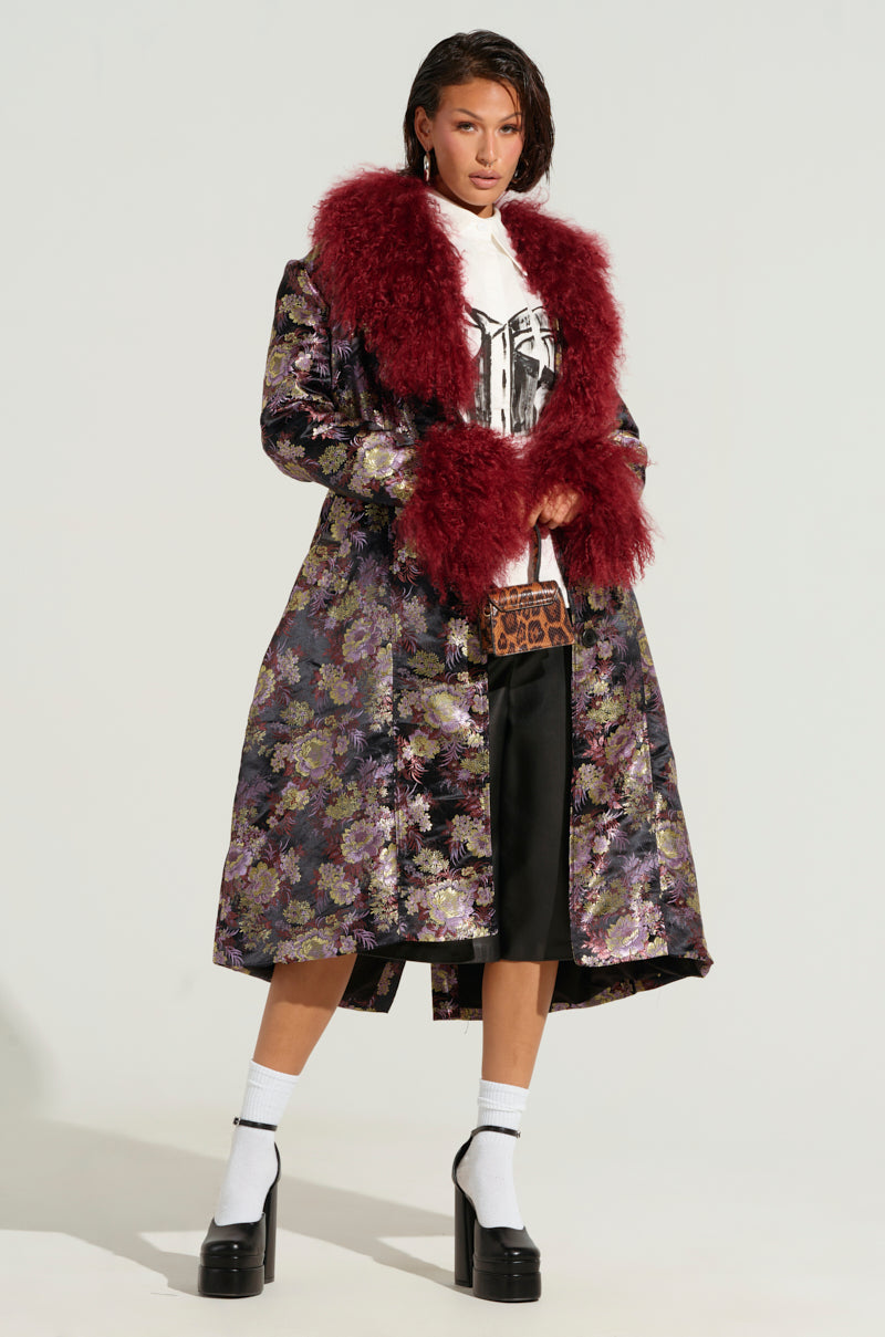 Dressed with confidence, the individual showcases the COSMO BROCADE TRENCH, easily distinguished by its vibrant faux fur lapel collar and floral brocade pattern. Complementing this standout piece is a white graphic t-shirt paired with black wide-leg pants. Their look is completed with white socks and black platform heels. The ensemble is accessorized by a small patterned bag, while their hair is slicked back for a sleek finish.