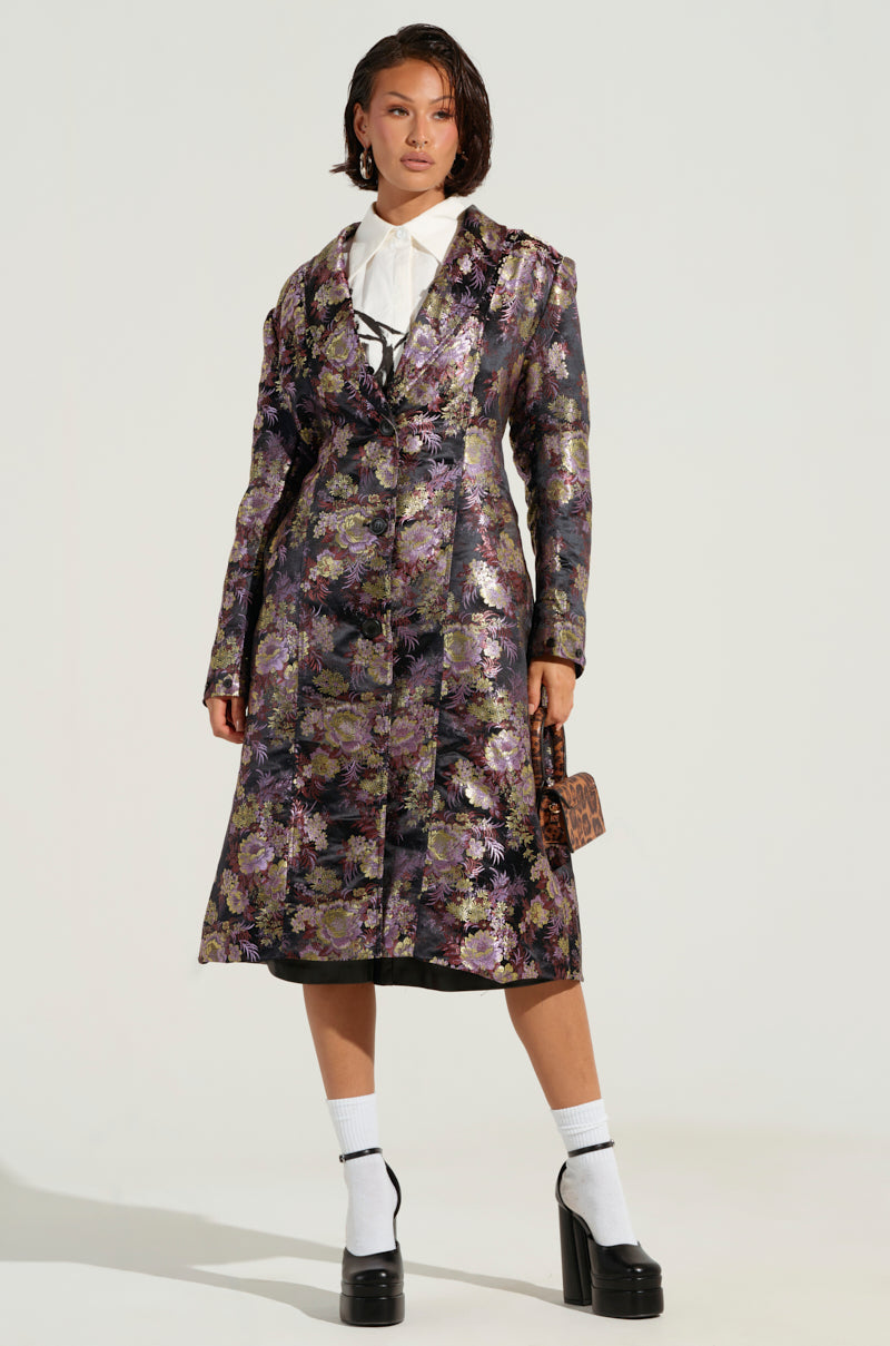 Wearing a COSMO BROCADE TRENCH, an individual exudes confidence as the long floral brocade design is accentuated by a faux fur lapel collar. They complement the ensemble with a small purse, black platform heels, and white socks, all set against a simple backdrop to highlight their bold fashion statement.