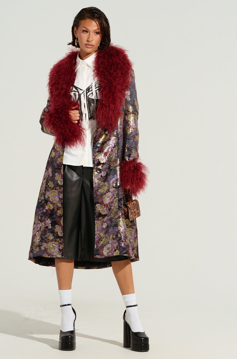 A person exudes confidence in a COSMO BROCADE TRENCH featuring a floral brocade pattern and a faux fur lapel collar, layered over a white hoodie. They pair this with knee-length black shorts, platform shoes, and carry a small leopard-print bag. The simple background accentuates the striking fashion ensemble.