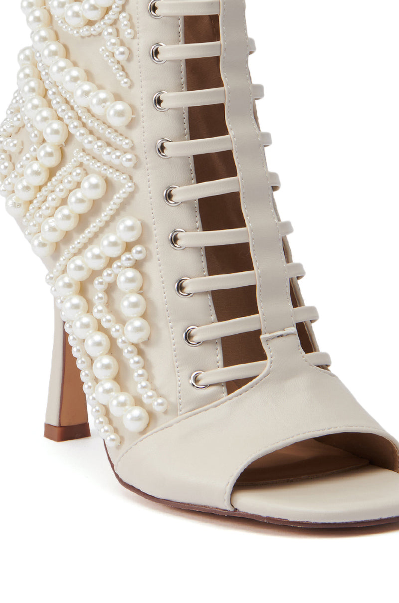 A close-up of the AZALEA WANG CHIPPER BONE BOOTIE WITH PEARL EMBELLISHMENT showcases its faux leather composition adorned with pearl embellishments and side lace-up detailing. This cream-colored beauty features an open-toe design with a slightly square heel, adding elegance to its unique style. 6