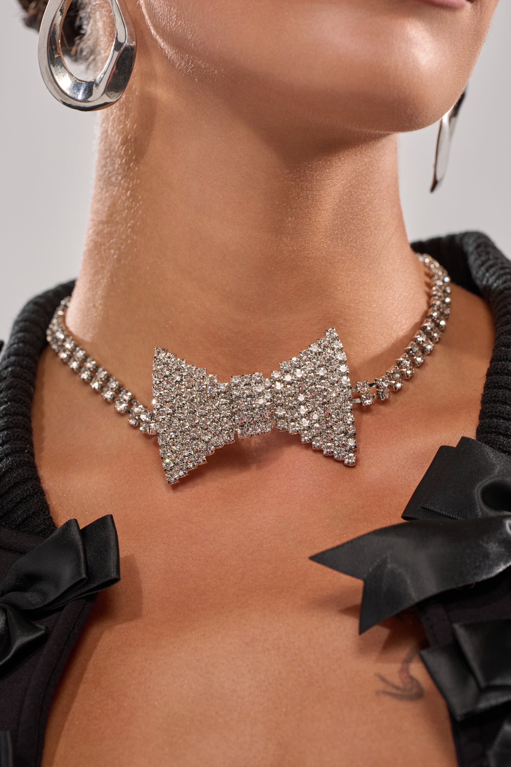 A woman shows off her lower face and neck, styled with the DAPPER CHOKER NECKLACE—a sparkling rhinestone chain with a bow pendant—paired with large silver hoops and a black top featuring satin bow details. 1