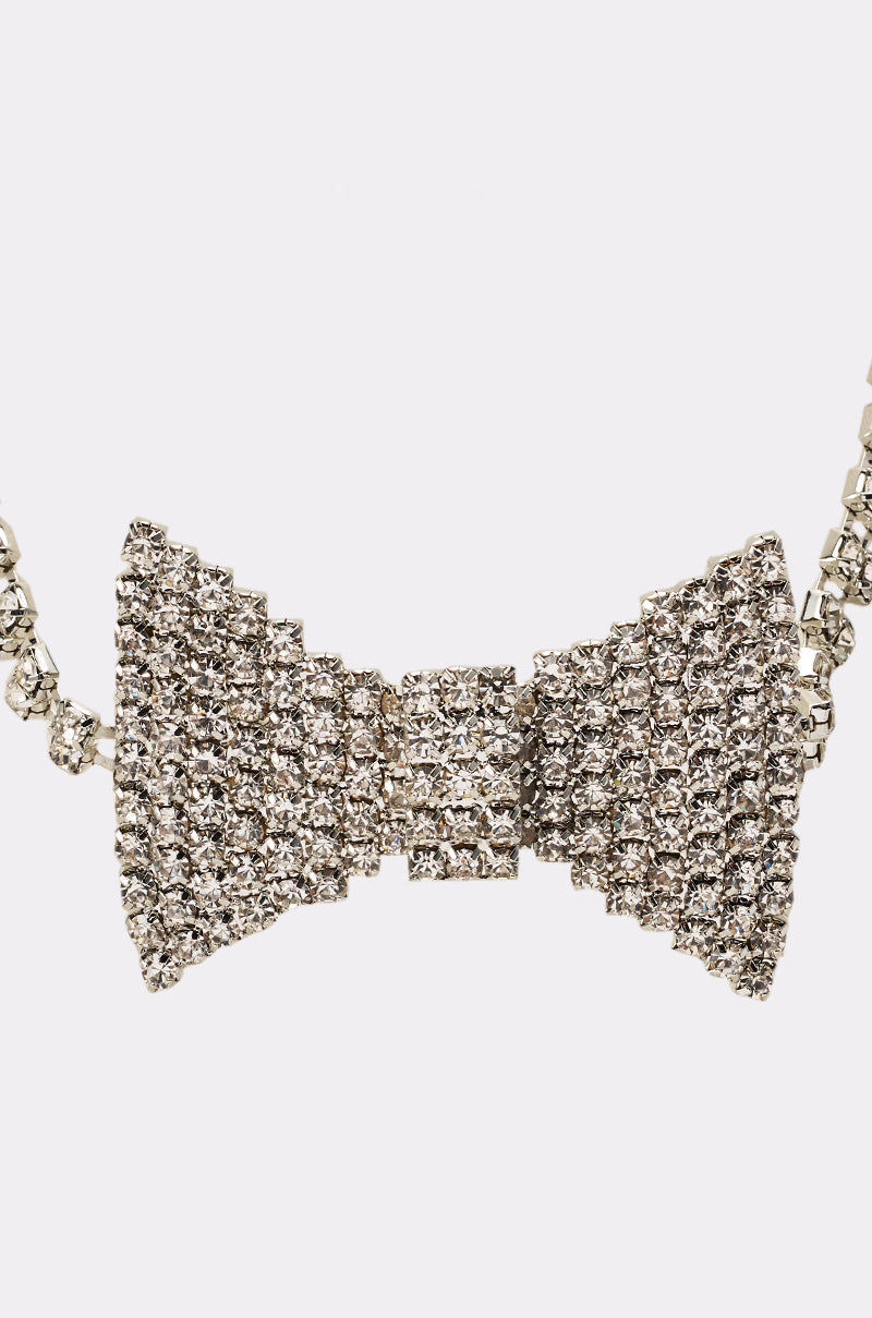 A close-up of the DAPPER CHOKER NECKLACE—a sparkling silver choker with a bow-shaped pendant adorned with shiny rhinestones—set against a plain, light background. 2
