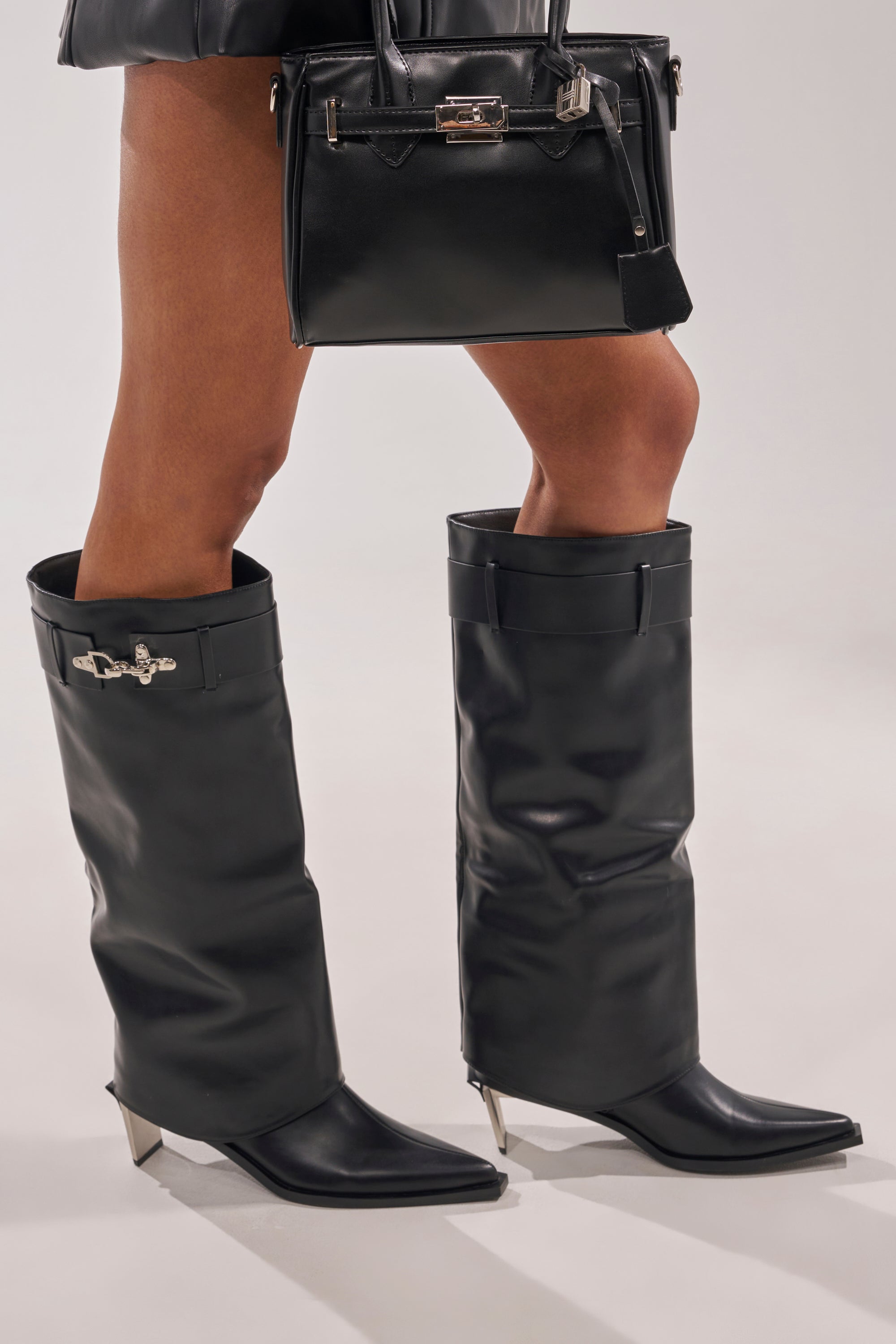 Wearing the AZALEA WANG CHRONO BLACK METAL HEEL BOOT—black faux leather, knee-high with pointed toes and a sleek geometric metal heel—paired with a black handbag, shown from mid-thigh down against a light background. 3