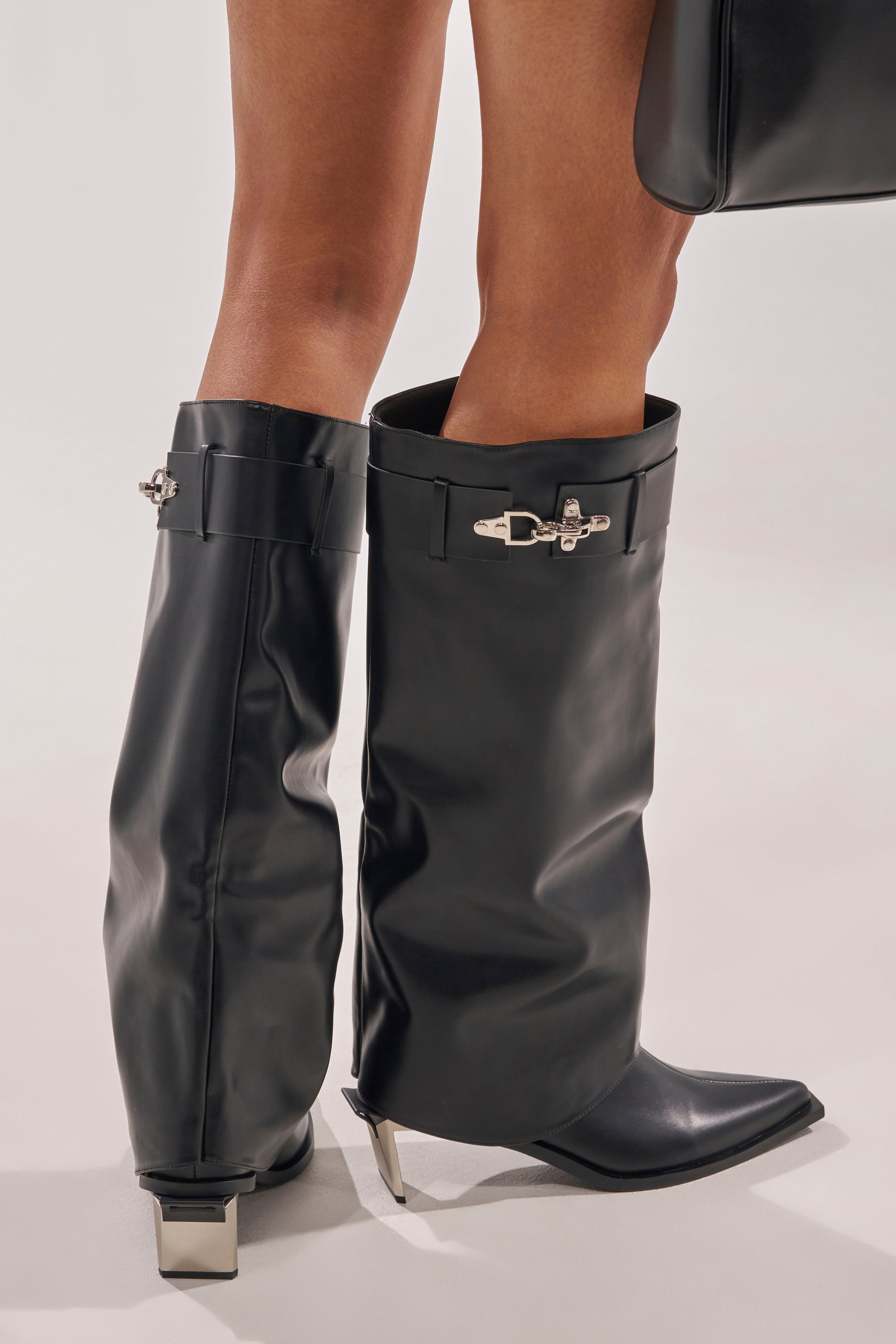 Someone stands on a light surface wearing the AZALEA WANG CHRONO BLACK METAL HEEL BOOT—oversized, black faux leather, knee-high boots with metallic hardware and a metal geometric heel—paired with a matching black bag. The boots are slouched below the knees. 8