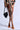 A person in a colorful, pattern-rich dress with intricate designs in brown, black, and beige stands against a neutral background. They hold a black faux leather handbag and wear AZALEA WANG CHUG ORANGE SANDALS.