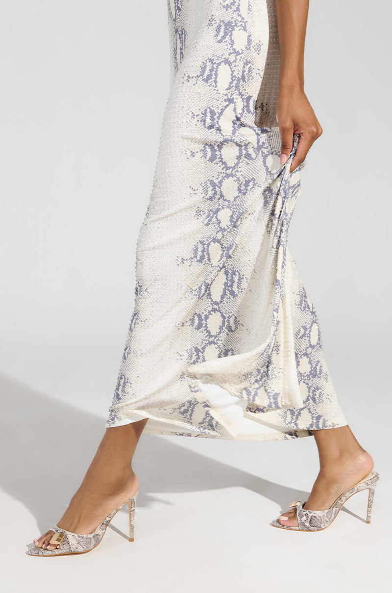 A person confidently struts in a full-length, white and gray snakeskin-patterned dress, perfectly paired with the elegant AZALEA WANG CHUG SNAKE SANDAL. The flowing design complements the high-heeled sandals as they gracefully walk, holding a portion of the dress in hand.