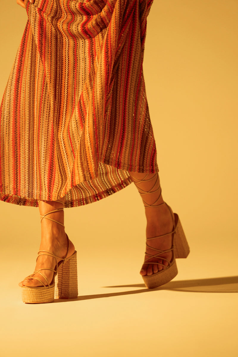 A person wears a colorful striped skirt and AZALEA WANG CIERRA BEIGE STRAPPY RAFFIA PLATFORM SANDALS with chunky heels, adding texture to the earth-toned outfit. The background glows with a warm, golden light.