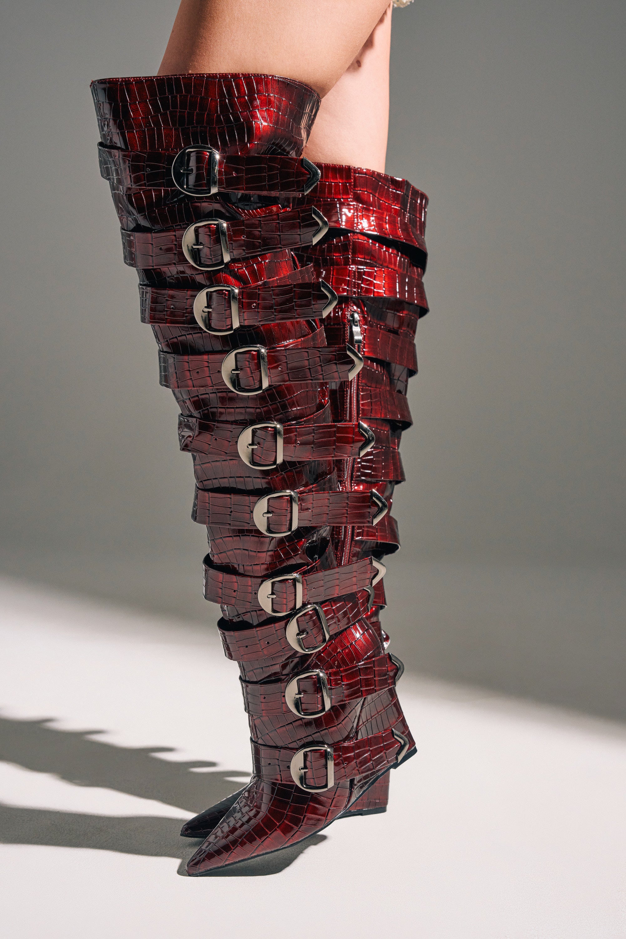 A person wears the AZALEA WANG CINZIA RED WEDGE BOOT—shiny, dark red faux crocodile thigh-highs with pointed toes and large silver side buckles, offering a bold, high-fashion statement.