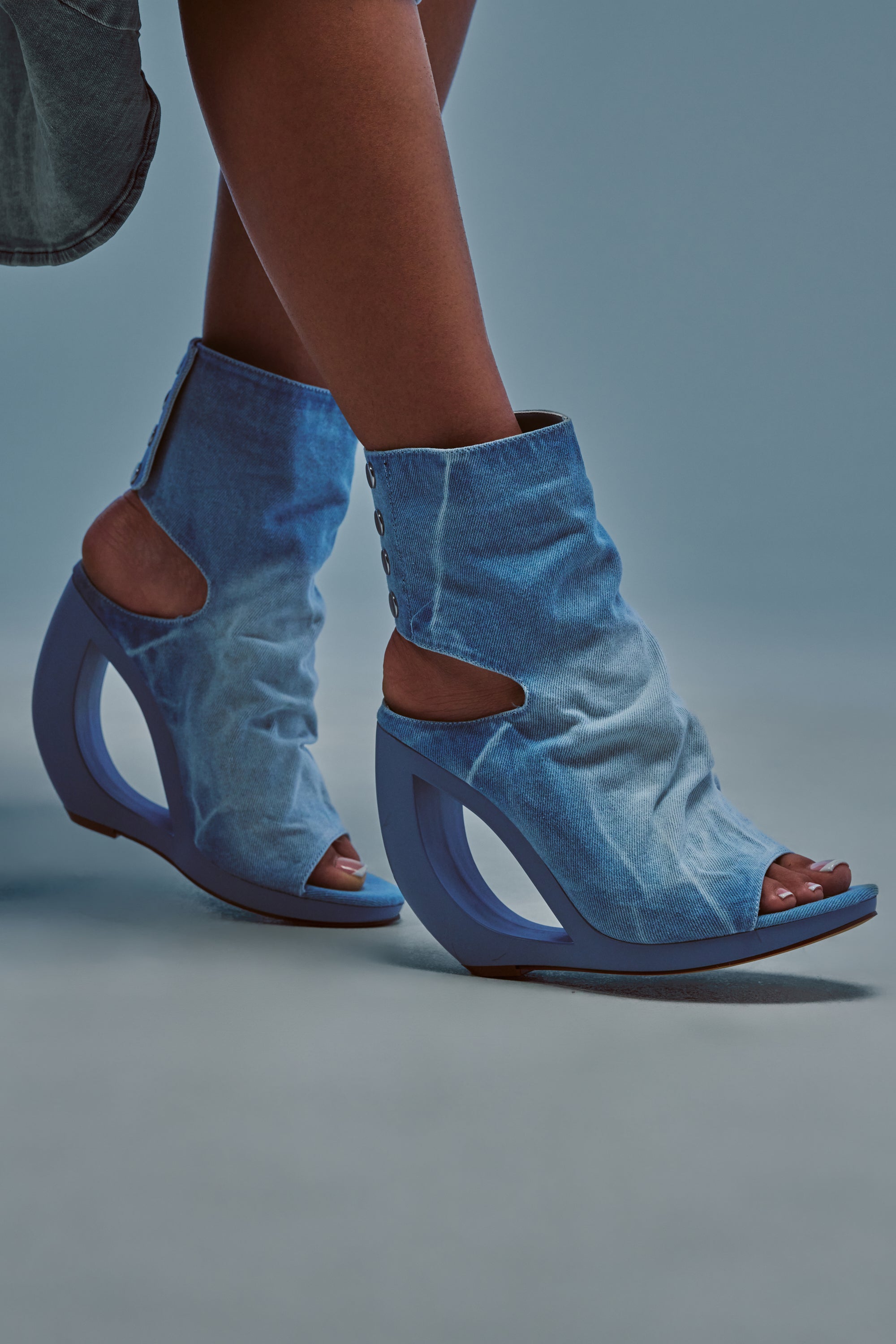 Wearing the AZALEA WANG CIRCOS LIGHT DENIM WEDGE BOOTIE—distinctive open-toe booties with curved, sculptural blue wedge heels—a person stands against a soft blue background, effortlessly blending summer streetwear style with bold footwear design.