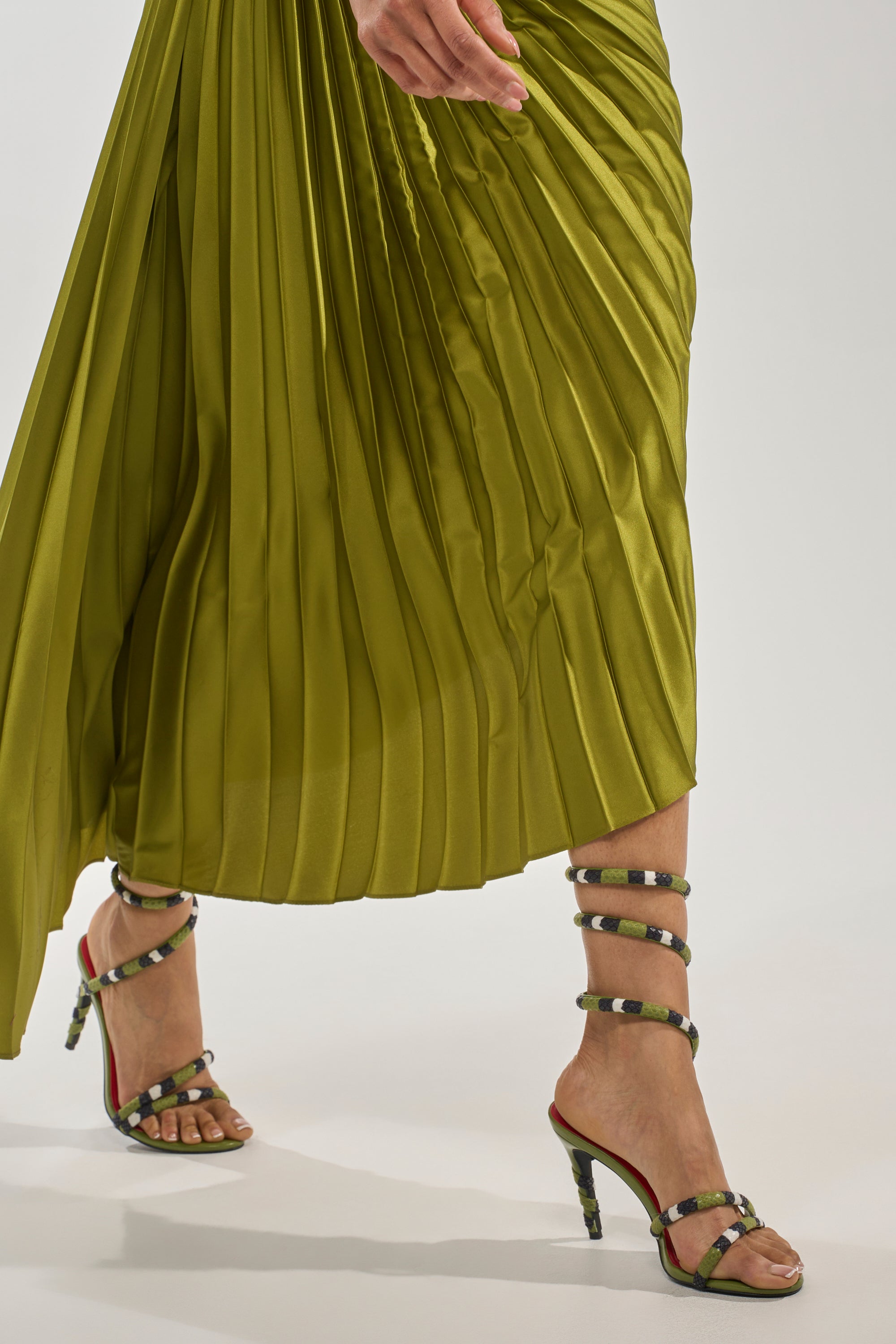 A person wearing an olive-green pleated skirt and AKIRA GOLD CITLALI GREEN SNAKE COIL SANDALS with snakeskin-textured straps walks on a plain, light-colored surface. 6