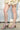 A person outdoors shows bare legs and feet in AZALEA WANG CLAREN BROWN MULTI WOODEN HEEL SANDALS, paired with a light ruffled skirt against a white wall and concrete ground.