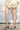 A person wearing a light, ruffled skirt and AZALEA WANG CLAREN BROWN MULTI WOODEN HEEL SANDALS stands on a concrete surface against a white wall.