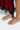 A close-up displays someone wearing the CLARISSA GOLD EMBELLISHED FLAT SANDAL, standing on a light surface. They're dressed in a red velvet dress with black lace trim elegantly flowing below the knees.