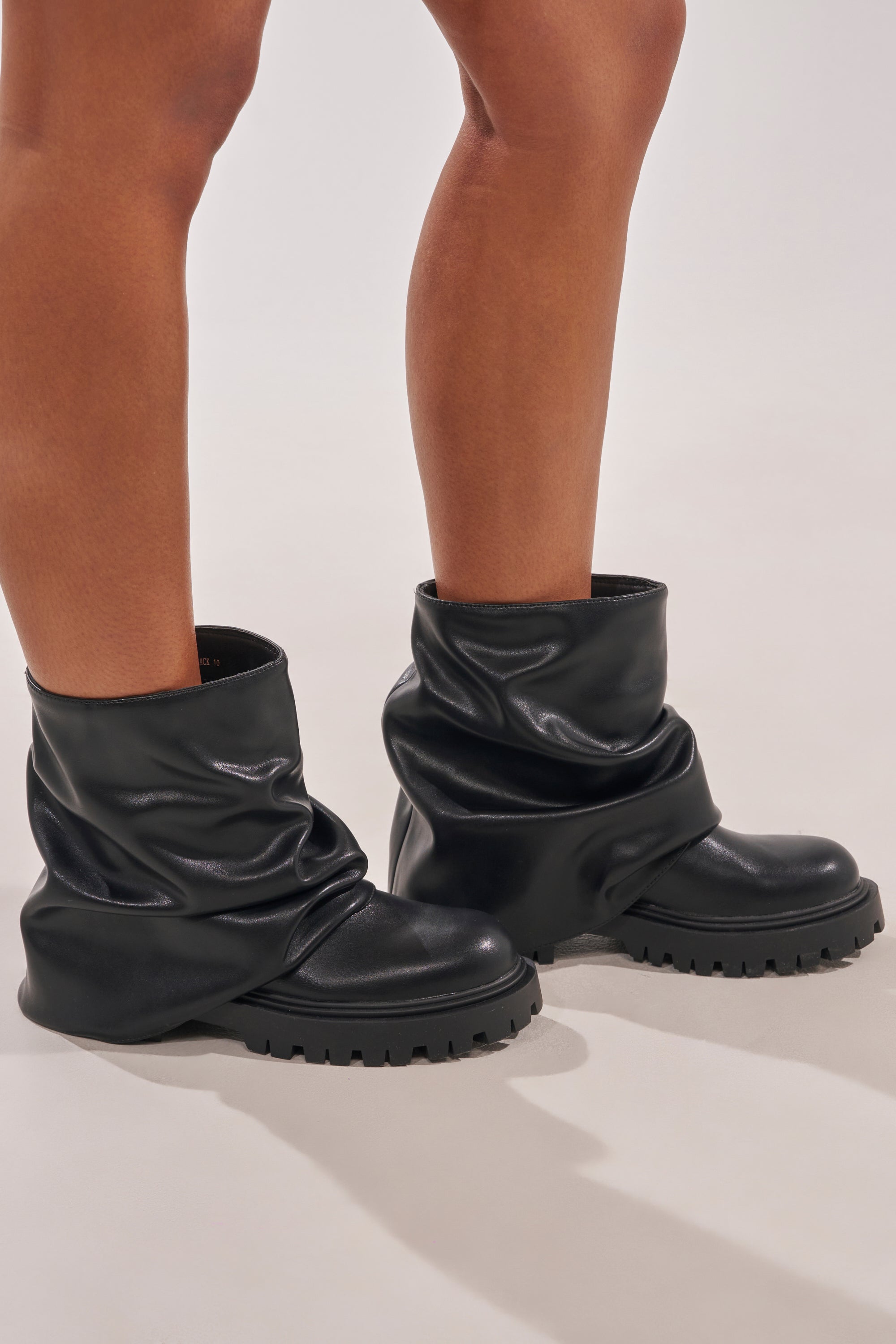 Only the lower legs and feet are visible of a person standing on a light-colored surface, wearing AZALEA WANG CLARKE BLACK PU SLOUCHY SHORT SHAFT COMBAT BOOTIES with chunky lug soles. 3