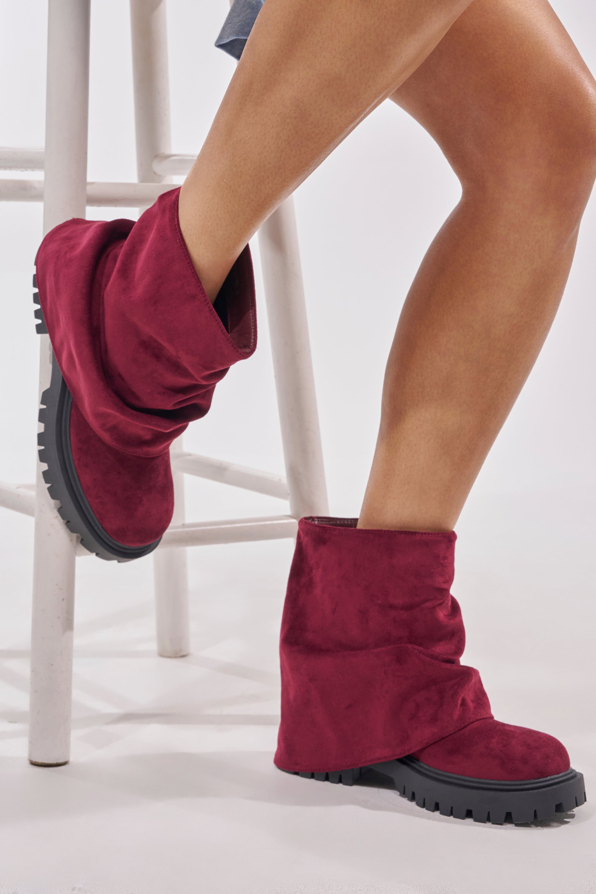 A person sits on a white stool, legs crossed, wearing the AZALEA WANG CLARKE FAUX SUEDE BURGUNDY FLAT BOOTIE with a relaxed foldover shaft and thick black soles for a laid-back, casual style.