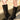 Someone wearing the AZALEA WANG CLEDQYN BLACK FAUX SUEDE BOOT with stiletto heels stands on gravel beside a concrete ledge, parked cars visible in the background.