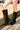 Someone wearing the AZALEA WANG CLEDQYN BLACK FAUX SUEDE BOOT with stiletto heels stands on gravel beside a concrete ledge, parked cars visible in the background.