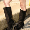 Someone wearing the AZALEA WANG CLEDQYN BLACK FAUX SUEDE BOOT with stiletto heels stands on gravel beside a concrete ledge, parked cars visible in the background.