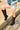A person stands outdoors in light denim cut-off shorts and AZALEA WANG CLEDQYN BLACK FAUX SUEDE BOOT, one foot on a concrete ledge, showcasing their outfit in bright sunlight.
