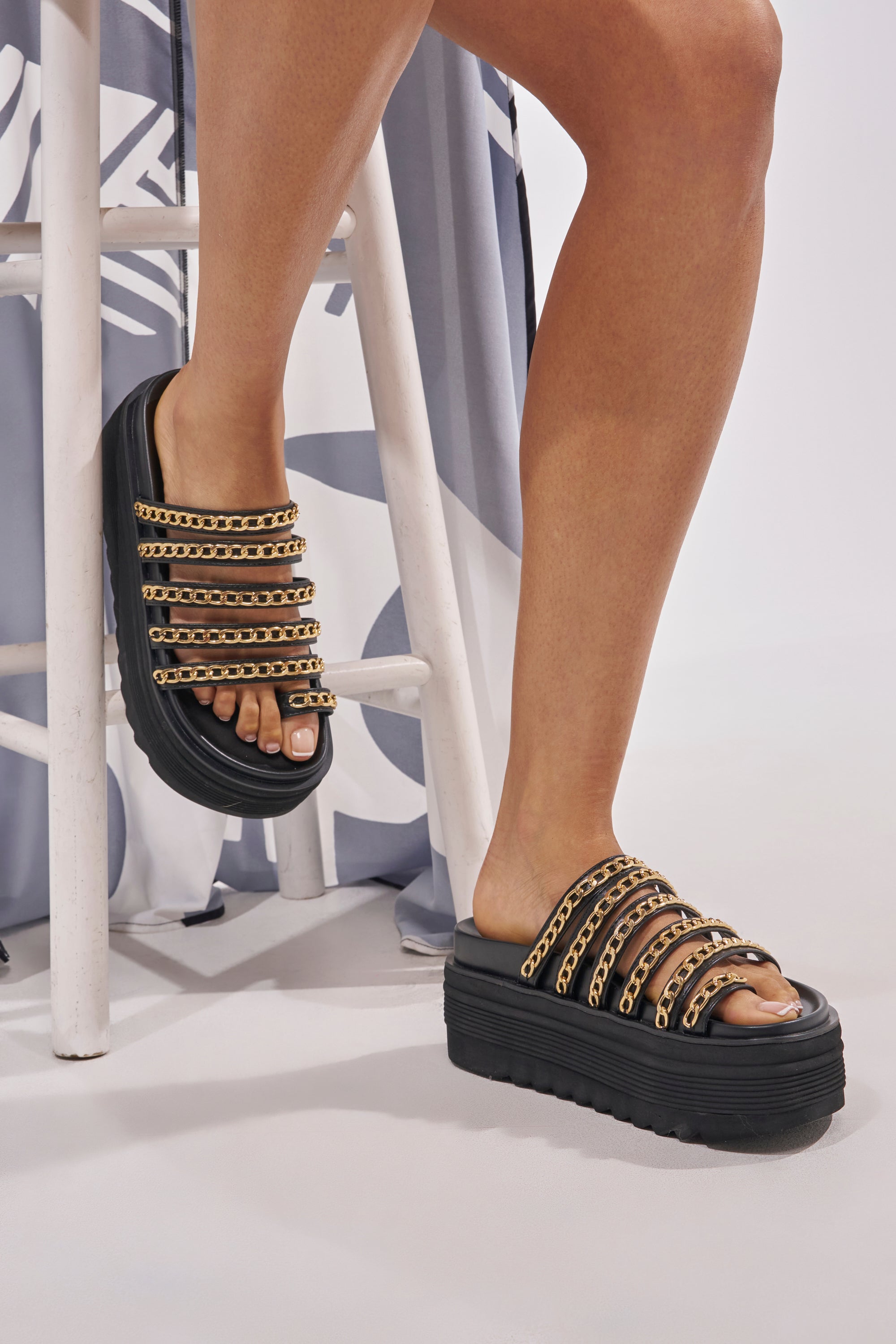Someone stands beside a white wooden stool on a patterned light gray and white floor, wearing the AKIRA CLEMENTE GOLD CHAIN DETAIL FLATFORM SANDAL in black faux leather. 0