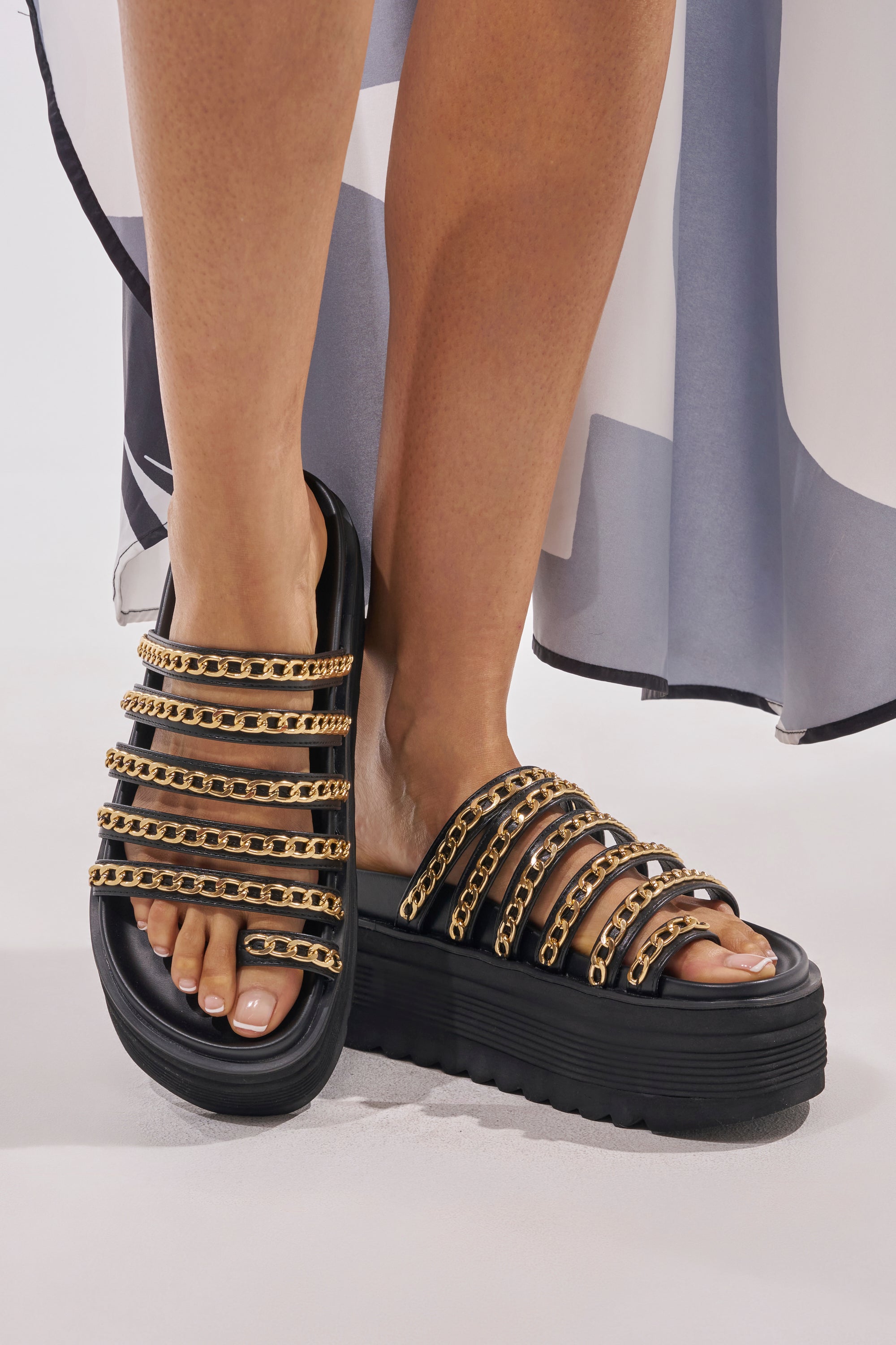 Someone stands on a light surface wearing the AKIRA CLEMENTE GOLD CHAIN DETAIL FLATFORM SANDAL in black with bold gold chain accents, paired with a flowy skirt featuring a geometric pattern. 2
