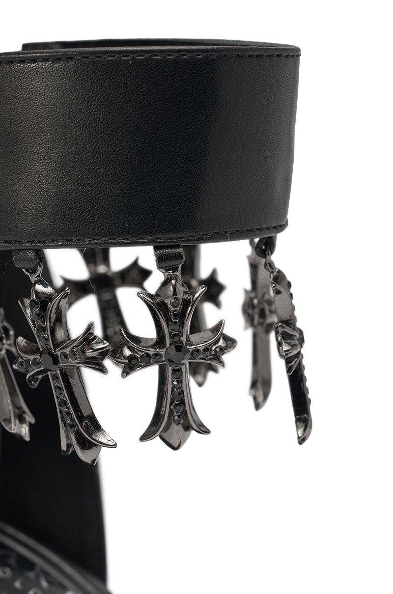 A close-up of the AKIRA GOLD CLERGY BLACK EMBELLISHED SEXY STRAP STILETTO HEEL showcases black leather and dangling rhinestone cross accents, highlighting its intricate, gothic-inspired design. 5