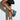 A person is posing against a white wall, wearing denim shorts and the chic AZALEA WANG CLIO DENIM SANDAL with a platform sole. They hold a black triangular handbag with large buckles, crossing their legs at the ankles to showcase the high wedge sandals with a denim design.