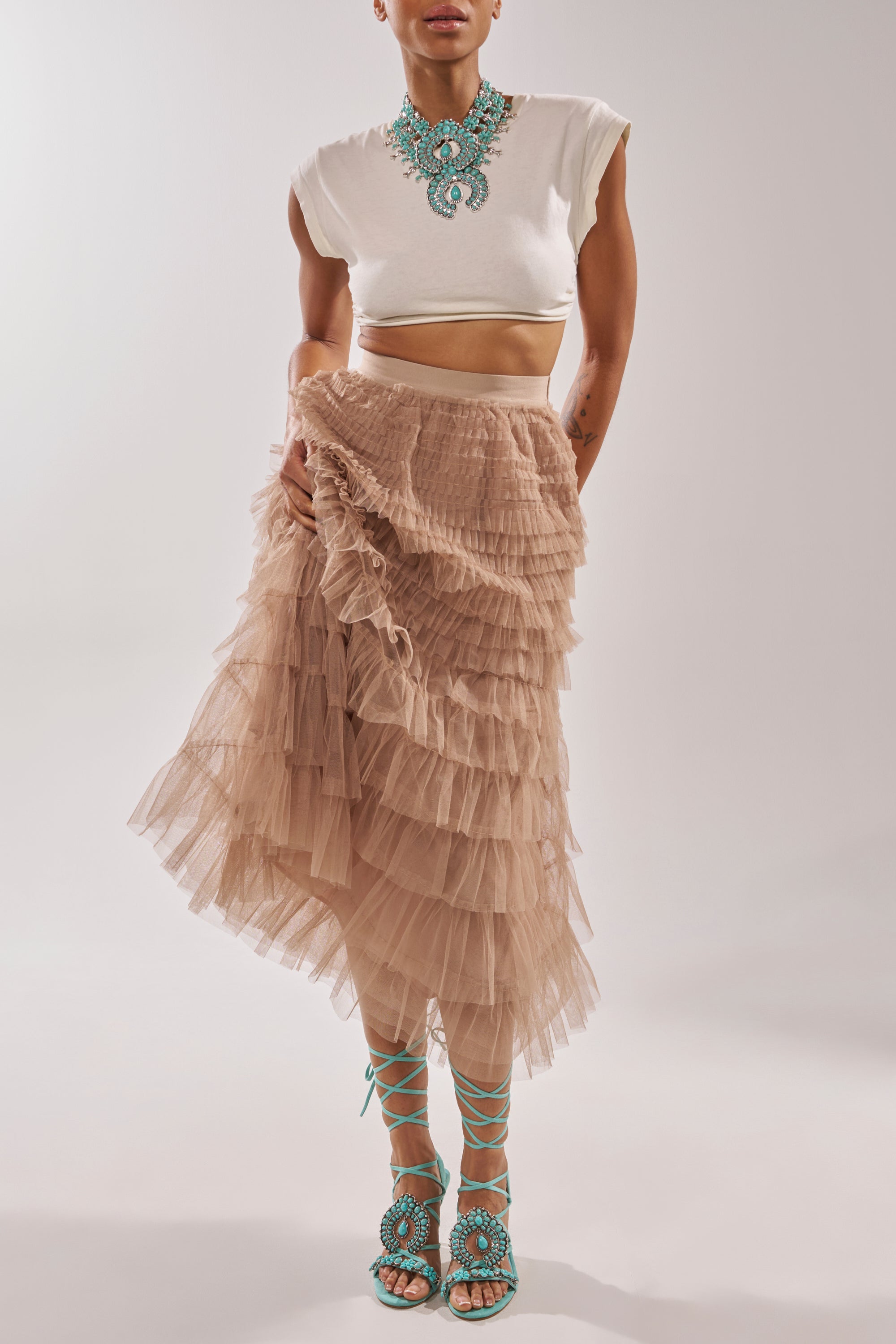 Person in a white cropped top, layered beige tulle skirt, AKIRA GOLD CLIPSE AQUA HARDWARE EMBELLISHED STILETTO SANDAL, and a large turquoise statement necklace stands against a plain light background. 2
