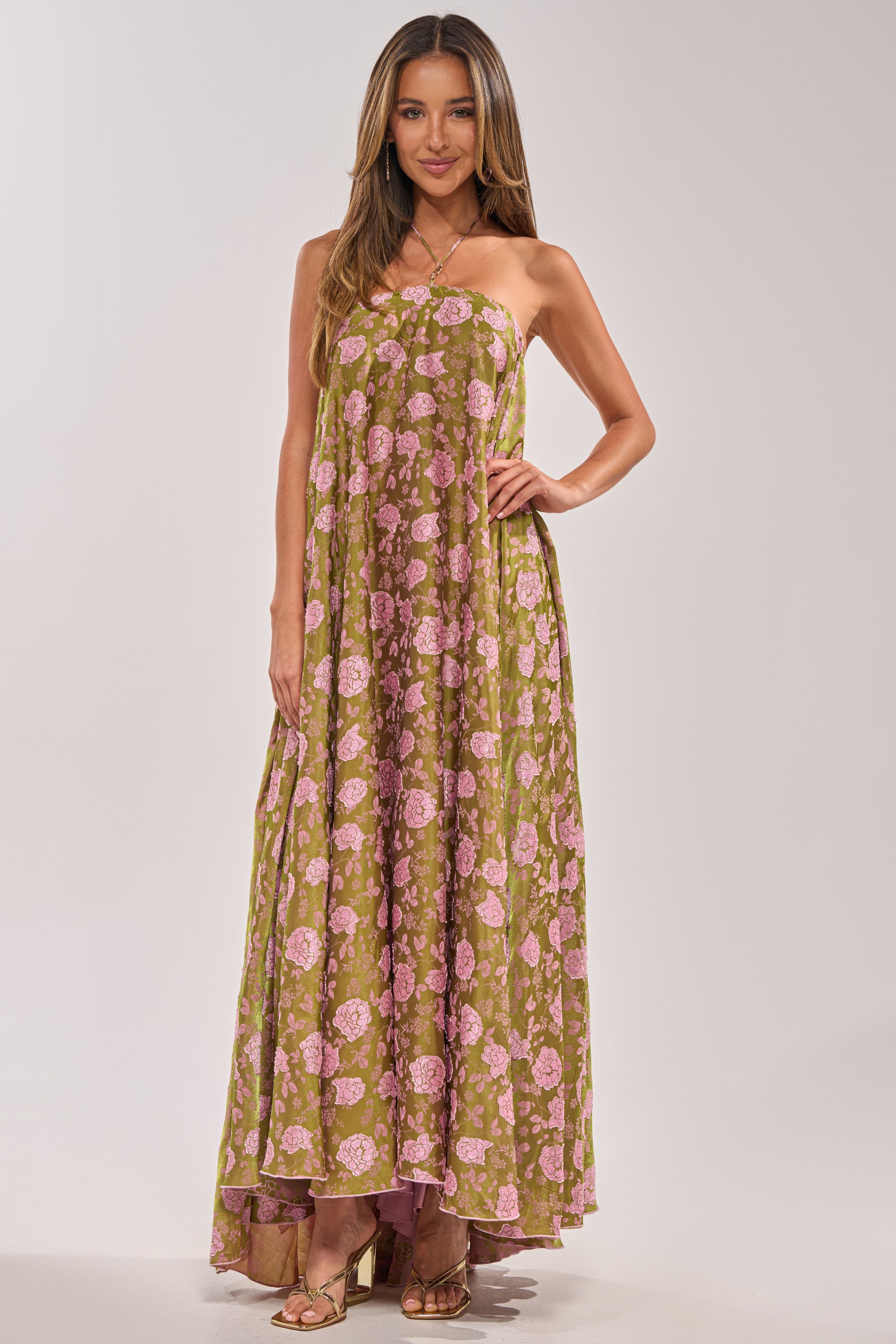 A woman wears the MIDNIGHT GARDEN FLORAL MAXI DRESS, a strapless green satin maxi featuring a pink floral print. She smiles gently with loose hair and beige heeled sandals—an elegant option for wedding guests.