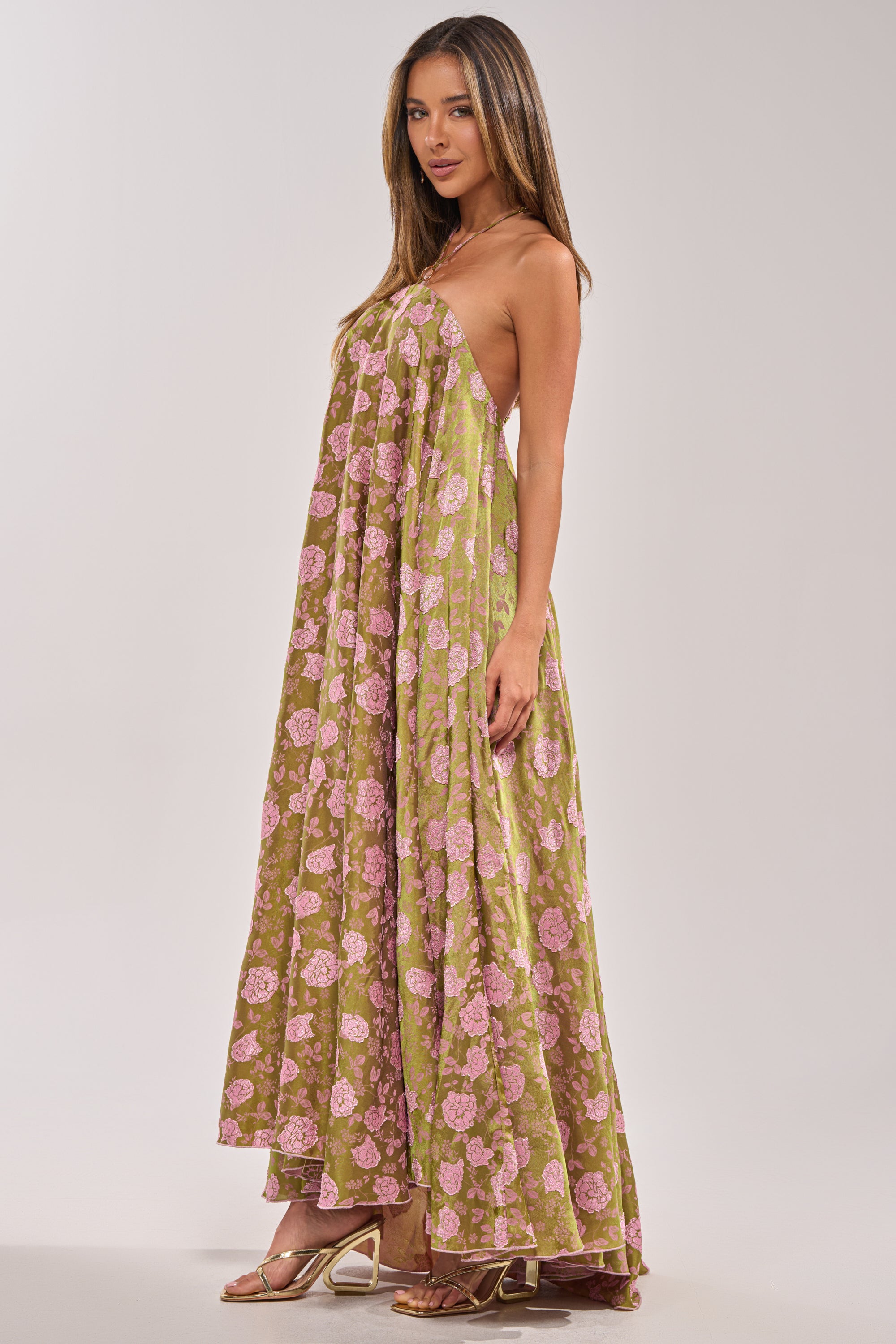 A woman poses in a studio wearing the MIDNIGHT GARDEN FLORAL MAXI DRESS, a flowy olive-green piece with floral embroidery and metallic open-toe heels. She faces sideways with long straight hair—ideal for a wedding guest look.