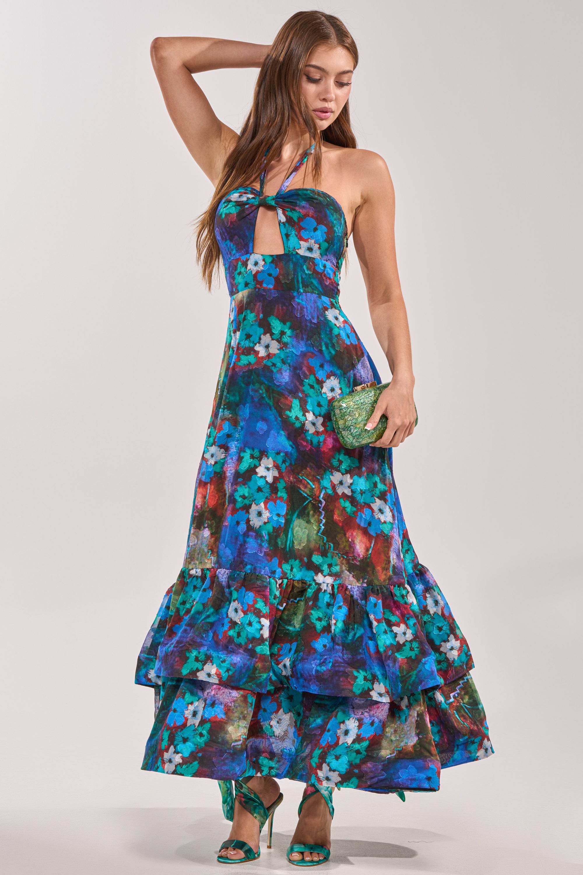 A woman models the HEADLINE WORTHY FLORAL MAXI DRESS, a sleeveless halter maxi dress with bold blue and green floral print—ideal for a wedding guest. She accessorizes with a green clutch, matching heels, and lets her long hair down.