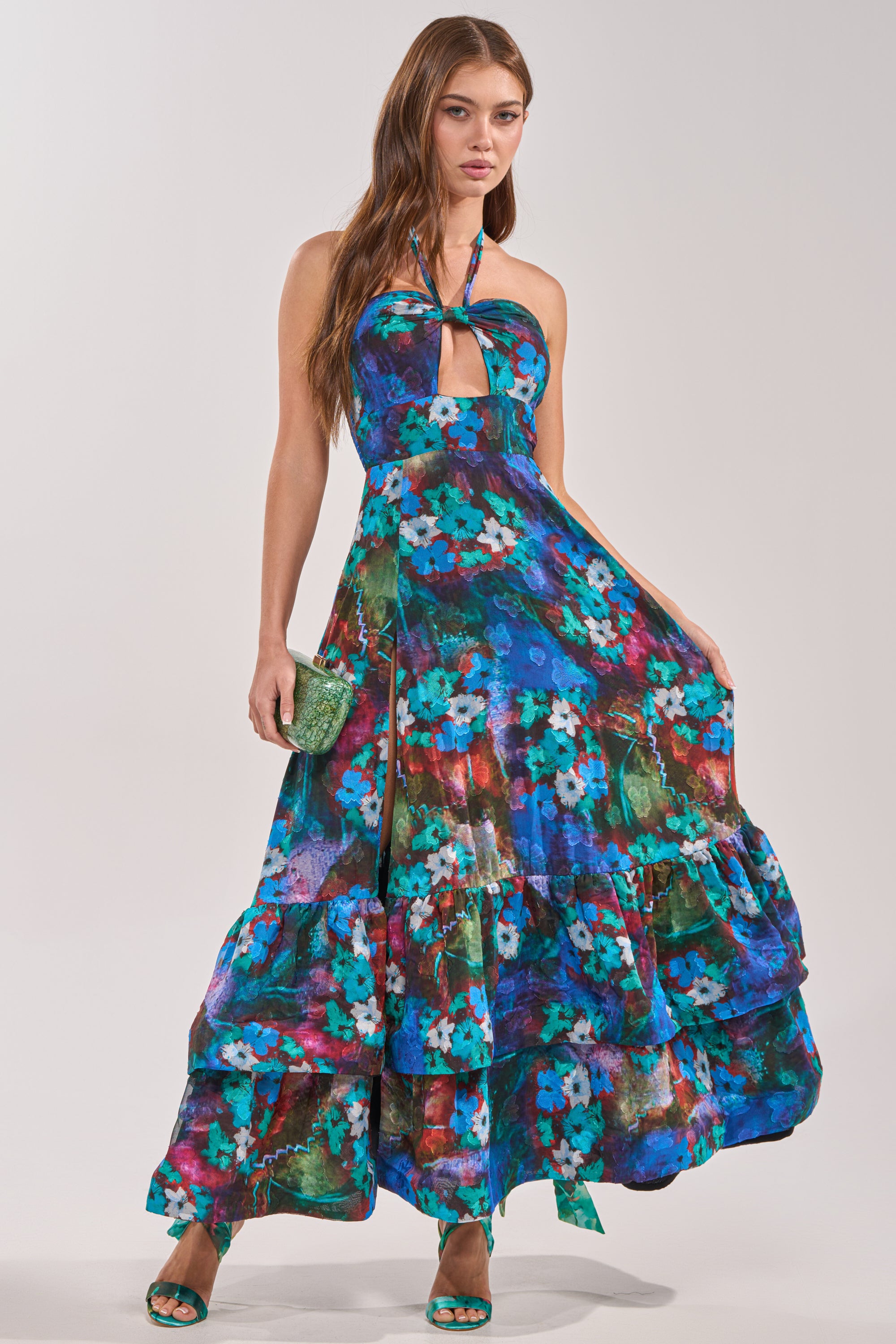 A woman wears the HEADLINE WORTHY FLORAL MAXI DRESS, a vibrant blue maxi with teal and magenta floral prints and a halter neckline. She pairs it with green heels and a green clutch—an ideal look for a wedding guest.
