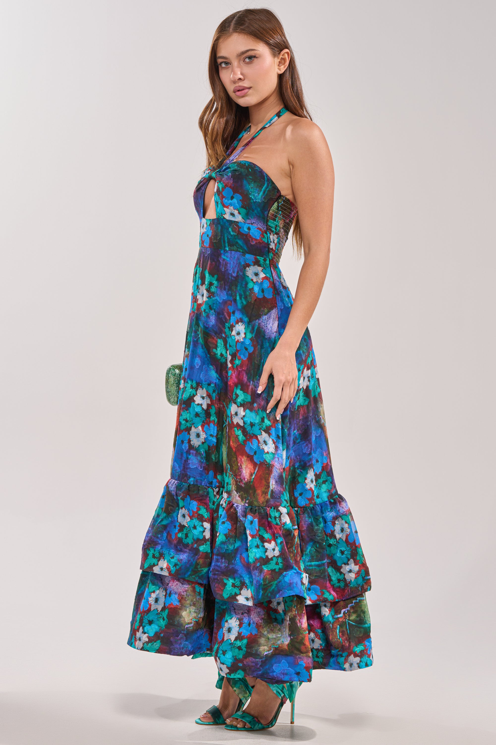 A woman models the HEADLINE WORTHY FLORAL MAXI DRESS, a blue halter gown with ruffled details and hem, teamed with green heels against a light background—an elegant option for wedding guests.