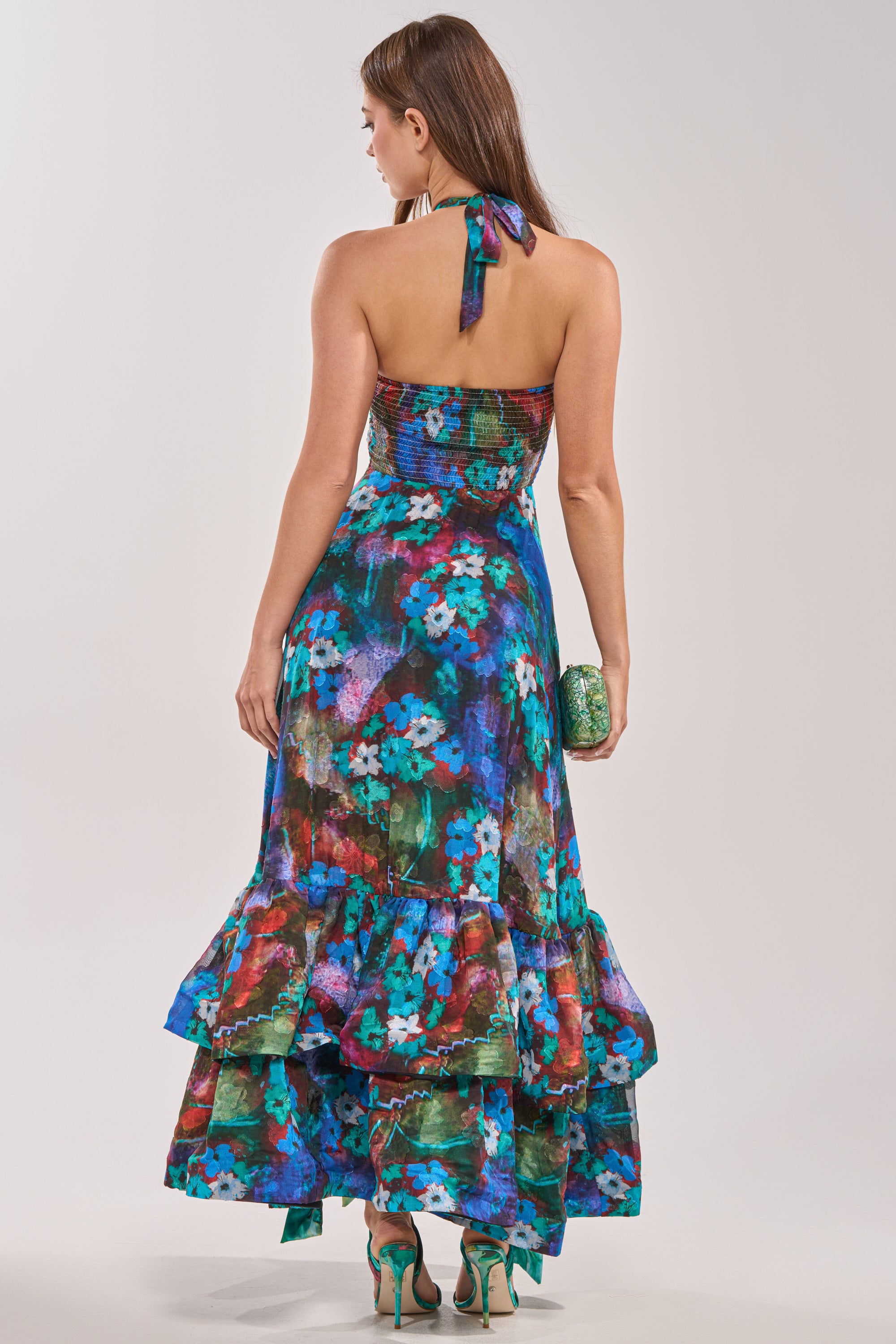 A woman with long brown hair wears the HEADLINE WORTHY FLORAL MAXI DRESS, featuring a colorful floral print and ruffled hem. She holds a green clutch and teal heels, shown from the back against a plain light background—perfect for weddings.