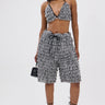 A woman stands against a plain background wearing SHAGGY BERMUDA SHORTS, a black and white textured crop top, strappy black heels, and holding a small black handbag.