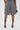A person stands facing away, wearing SHAGGY BERMUDA SHORTS made from carpet-like fabric, black lace-up high-heeled sandals, and holding a small black handbag against a plain white background.