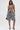 A woman stands against a plain white background wearing the SHAGGY BRALETTE and matching knee-length shorts, holding a small black handbag and wearing black strappy high-heeled sandals.