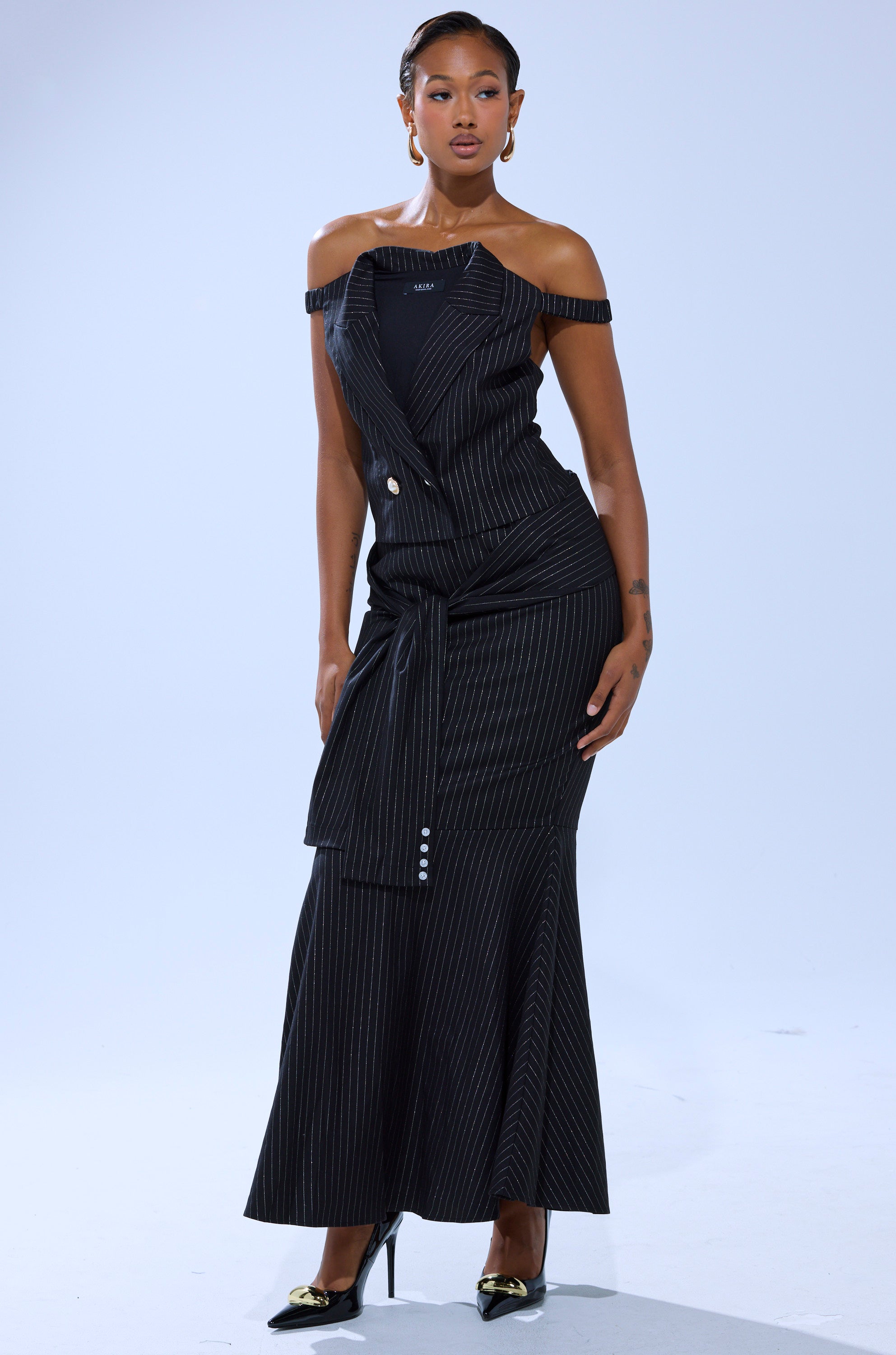 A woman models the BUSINESS ONLY PINSTRIPE CORSET, a black pinstripe dress with a belted waist and long skirt, styled with black heels featuring gold accents and hoop earrings for a modern business chic look against a plain light background.