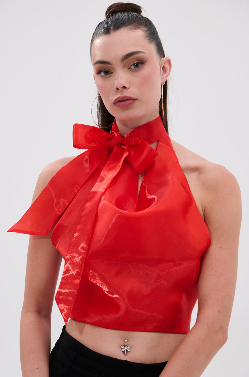 A woman with long brown hair in a high ponytail wears the SUNSHINE COWL NECK SATIN BLOUSE IN ORANGE, styled as a halter crop top with a large neck bow. She accessorizes with hoop earrings and a butterfly navel piercing against a plain white background.