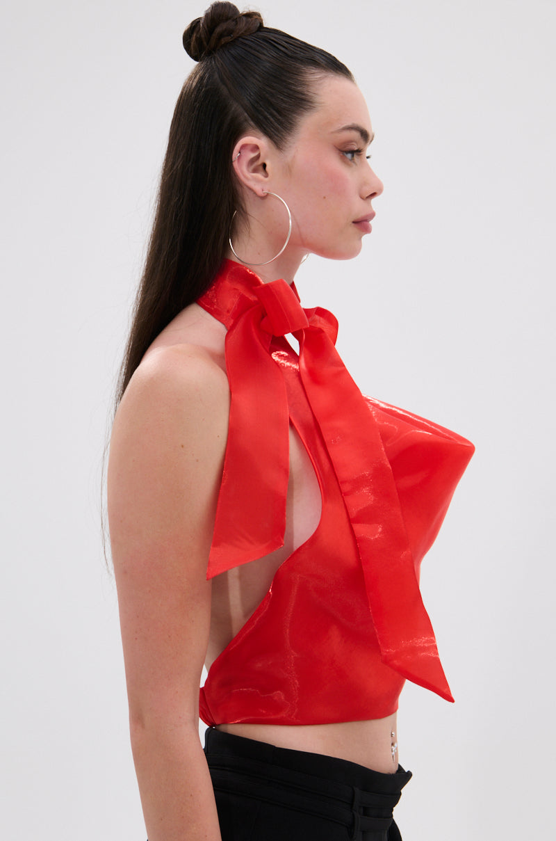 A woman with long dark hair in a top knot wears the SUNSHINE COWL NECK SATIN BLOUSE IN ORANGE, styled with large hoop earrings and black pants, posed in profile against a plain background.
