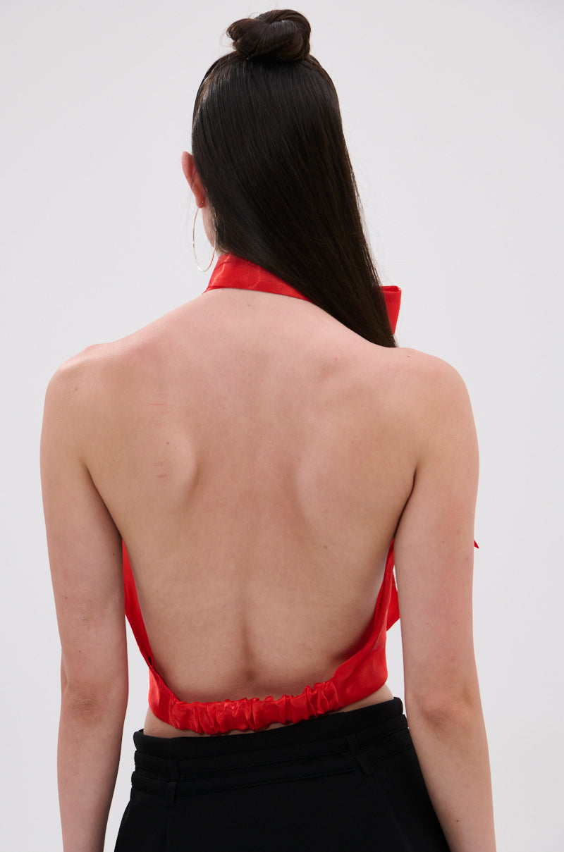 A person with long dark hair in a top bun is seen from behind wearing a backless Sunshine Cowl Neck Satin Blouse in Orange and black pants against a light gray background.