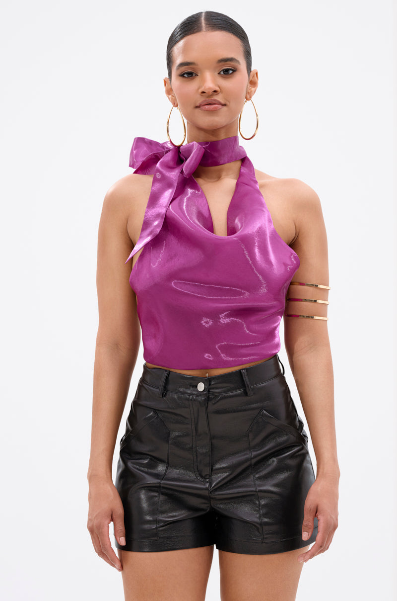 A woman in the SUNSHINE COWL NECK SATIN BLOUSE IN PURPLE, featuring a shiny cowl neck, open-back with a large bow, and paired with black high-waisted leather shorts and bold accessories, stands against a plain white background. 1